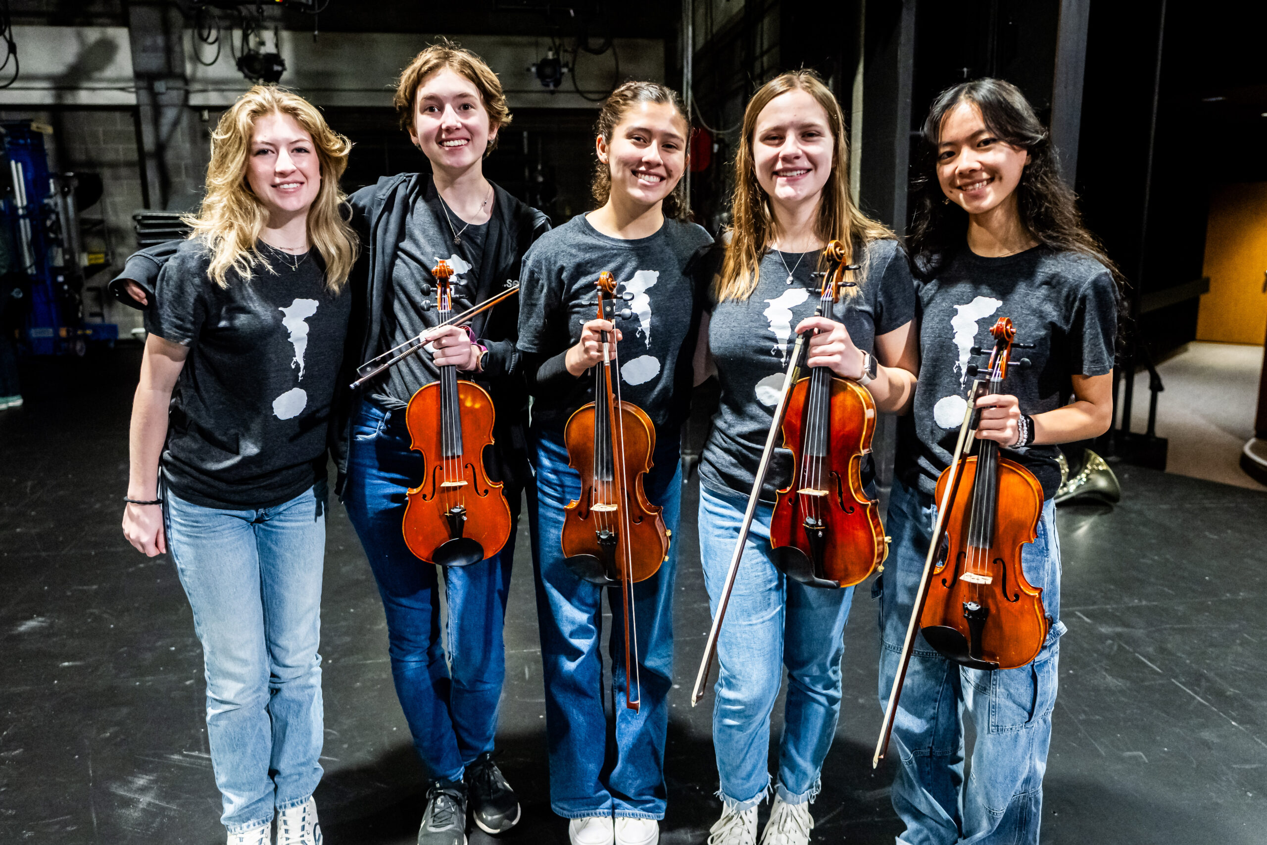 The image includes five smiling student musicians who are holding violins and have their arms around each other in a friendly gesture